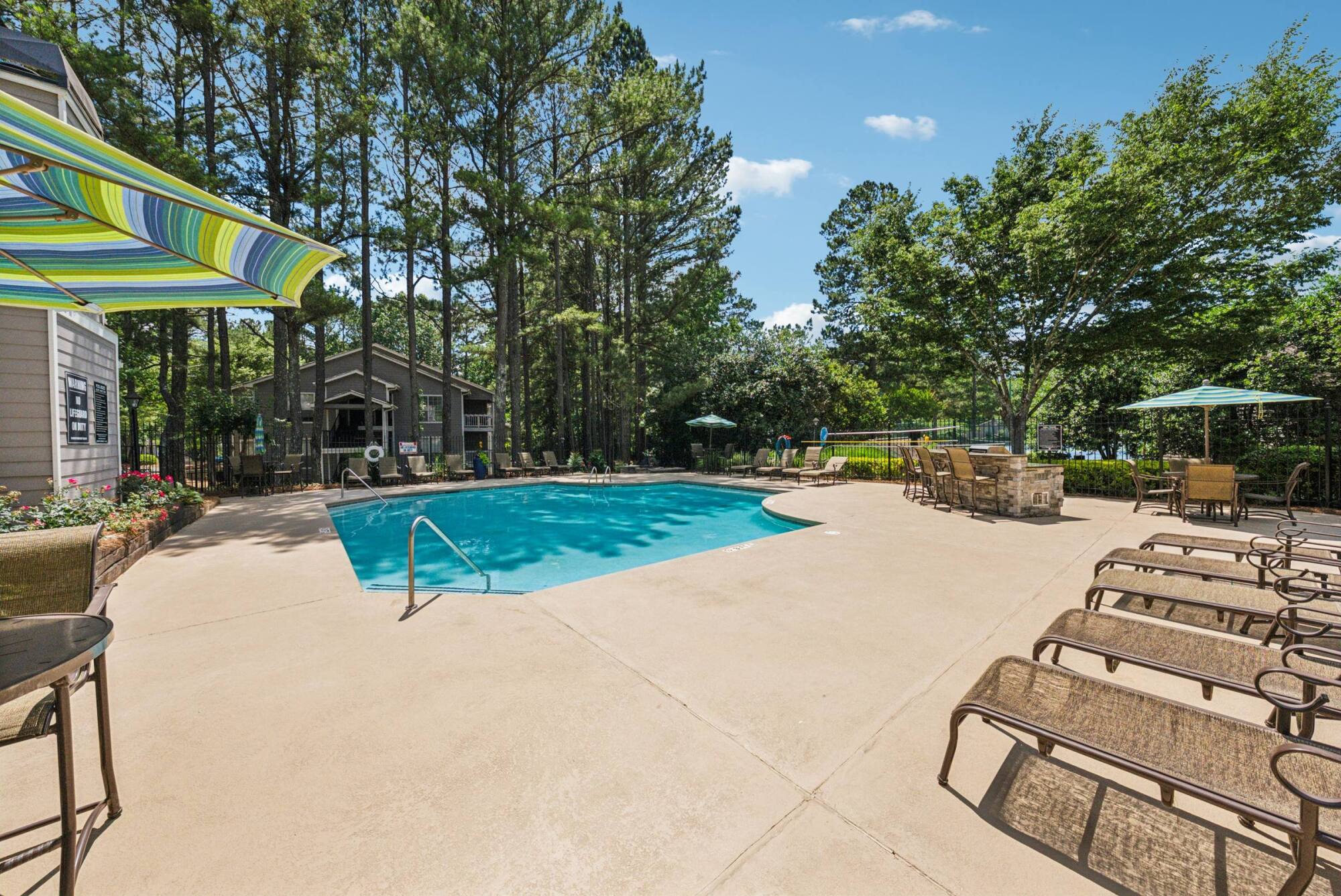 Outdoor swimming pool surrounded by lounge chairs, tables, and trees under a clear blue sky.