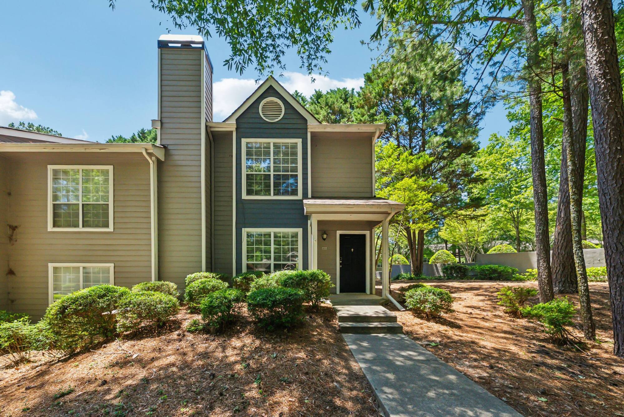Two-story gray house with large windows and a front porch, surrounded by trees and shrubs on a sunny day.